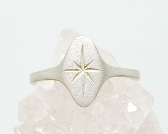 May include: A silver ring with an oval face featuring an engraved eight-pointed star design. The ring is set against a light background, highlighting the ring's details and shape. The ring is a piece of jewellery.