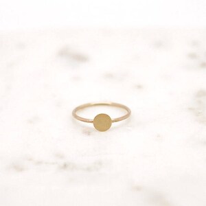 May include: A delicate gold ring with a small, circular design. The ring is set against a white background, highlighting its simple elegance. The ring's band is thin, and the circular design is centered. A minimalist piece of jewelry.
