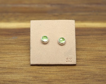 May include: A pair of stud earrings with round, light green gemstones set in silver. The earrings are displayed on a small, square, tan leather card. The card has the initials "S S" stamped on it.