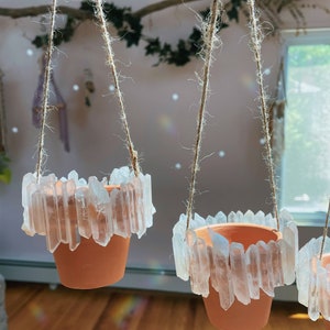 May include: Three terracotta pots hanging from twine, each with a rim of clear quartz crystals. The pots are hanging in a room with a light pink wall and a window in the background.