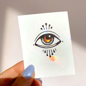 May include: A sticker with a black and white illustration of an eye with an orange and yellow iris. The eye is surrounded by a circle of rays.