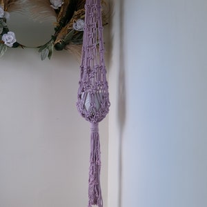 May include: A lavender macrame plant hanger with a long tassel. The hanger is hanging from a white wall.