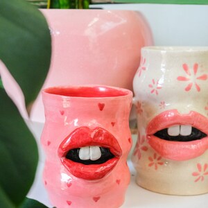 May include: Two ceramic mugs with pink and white glaze. One mug has a pink heart pattern and a red mouth with white teeth. The other mug has a white background with pink flower patterns and a pink mouth with white teeth.