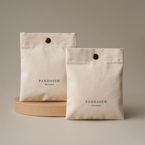 May include: Two off-white canvas pouches with a snap closure and the text "PANDASEW PACKAGE" printed in black. The pouches are on a small, round, light brown platform. The background is a neutral beige.