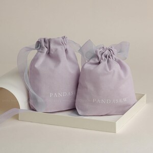 Peut inclure: Deux sacs à cordon de couleur lavande avec des rubans blancs. Le plus grand sac porte l'inscription "PANDASEW" imprimée sur le devant. Le plus petit sac porte l'inscription "PANDASE" imprimée sur le devant.