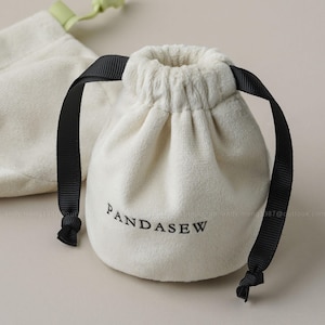 Puede incluir: Bolsa de terciopelo color crema con lazos negros. La bolsa tiene la palabra "PANDASEW" impresa en negro. Otra bolsa es visible en el fondo.