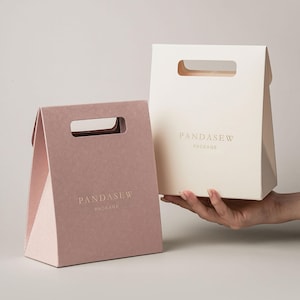 Pode incluir: Duas sacolas de presente de papel triangulares com alças integradas. Uma sacola é rosa seco e a outra é creme. Ambas apresentam o texto dourado PANDASEW PACKAGE impresso na frente. As sacolas têm um acabamento fosco e um design dobrado.