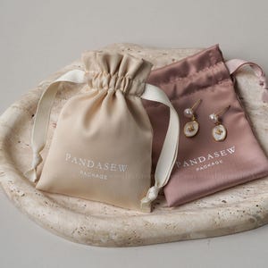 May include: Two satin drawstring pouches, one beige and one mauve, with the text "PANDASEW PACKAGE" printed on them. A pair of gold earrings with pearls and white oval pendants are next to the mauve pouch. The pouches are on a textured, light-colored surface.