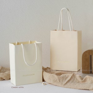 May include: Two rectangular paper gift bags with white rope handles. The shorter bag is cream white and the taller bag is beige. Both bags feature the text PANDASEW printed in gold on the front. The bags are positioned on a textured fabric surface.
