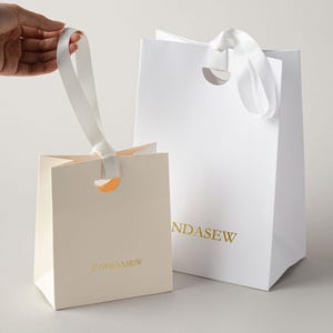 Puede incluir: Una bolsa de regalo pequeña de papel color crema y una bolsa de regalo más grande de papel blanco, ambas con el texto en lámina dorada que dice PANDASEW. Cada bolsa tiene una abertura troquelada con una cinta de satén blanca para llevar.