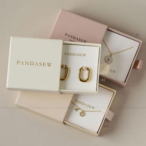 Pode incluir: Uma variedade de caixas de joias em tons de branco e rosa, cada uma contendo brincos e colares dourados. As caixas são rotuladas com a marca "PANDASEW" em letras douradas. As joias incluem um colar com a letra "L" e outro com a letra "H".