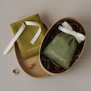 May include: Two small olive green and forest green velvet pouches with white ribbon ties, one inside a wooden oval container. The pouches have the word "PANDASEW" printed on them. A small gold-colored ring and a gold-colored earring are also visible.
