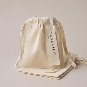 May include: A beige cotton drawstring pouch featuring a vertical fabric tag printed with the text PANDASEW PACKAGE in black ink. The pouch has a textured, natural fibre appearance with matching woven drawstrings on both sides of the opening.
