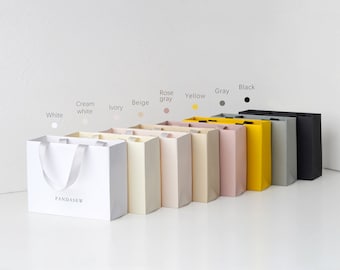 Puede incluir: Un conjunto de diez bolsas de regalo de papel en varios colores, incluyendo blanco, crema, marfil, beige, gris rosado, amarillo, gris y negro. Las bolsas tienen asas y están etiquetadas con el nombre de la marca "PANDASEW".