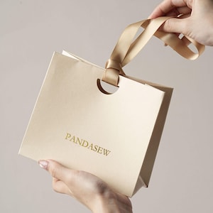 May include: A light beige paper gift bag featuring the word PANDASEW printed in gold lettering on the front. The bag includes a circular cutout at the top centre and a matching light beige satin ribbon handle attached through the opening.