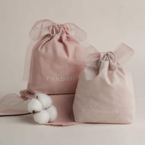 Puede incluir: Dos bolsas rosas con cordón y detalles de cinta transparente. Las bolsas tienen bordado el texto "PANDASEW". Una ramita de algodón añade un toque suave. Las bolsas están hechas de tela suave.