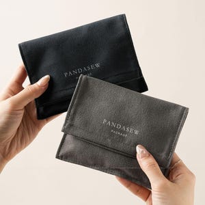 Pode incluir: Duas bolsas retangulares de textura macia nas cores preta e cinza-escura. Cada bolsa possui um fecho de aba e o texto impresso em branco PANDASEW PACKAGE centralizado na parte frontal. As bolsas são seguradas exibindo suas superfícies frontais.