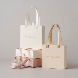 May include: Cream-colored gift bags and boxes with satin ribbon handles. The bags and boxes are stacked, with the brand name "PANDASEW" printed in gold. The boxes are in cream and pink.