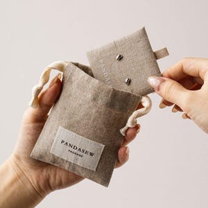 Pode incluir: Uma bolsa de linho bege com cordão e uma etiqueta de tecido branco com a inscrição PANDASEW PACKAGE. Um cartão de linho bege a condizer é parcialmente retirado da bolsa, com dois pequenos brincos prateados em forma de feijão na superfície.