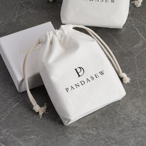 Puede incluir: Una bolsa de algodón blanco con cordón de ajuste, con un logotipo "P" negro y el texto "PANDASEW" impreso en la parte delantera.