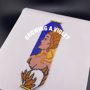 May include: A beaded bracelet featuring a colourful image of a woman wearing a crown. The bracelet has gold-coloured clasps and the text "GROWING A VIOLET" is printed on the bracelet.