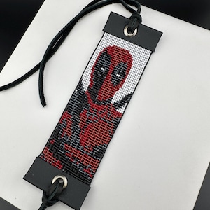 May include: A handmade bookmark with a pixelated Deadpool design in red, black, and white. It has black leather ends with silver eyelets and a black leather cord. A unique gift for comic book fans. The bookmark is approximately 18 cm in length.