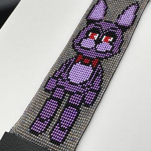 Könnte beinhalten: Lila und schwarzes Perlenarmband mit einem Pixelbild eines Comic-Charakters mit roten Augen und einer Schleife.