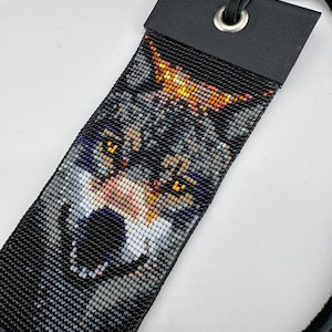 May include: A black leather bookmark with a beaded wolf design. The wolf's eyes are yellow and the beadwork is detailed.