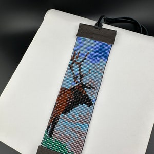 May include: A beaded bracelet with a brown elk design against a blue and green background. The bracelet has brown leather ends with a black cord.
