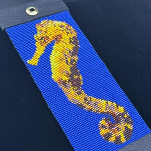 May include: A beaded bracelet with a seahorse design. The seahorse is in yellow, brown, and orange against a blue background. The bracelet has black leather ends with a metal grommet. The bracelet is approximately 20 cm long.