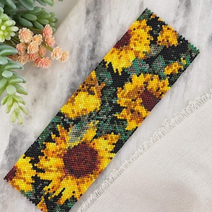 Op de afbeelding: Een kralenarmband met een zonnebloemmotief. De armband toont gele zonnebloemen met bruine centra en groene bladeren op een zwarte achtergrond. De rechthoekige armband is gemaakt van kleine kralen en toont een gedetailleerd bloemenontwerp.