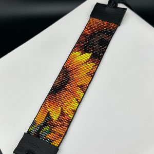May include: A beaded bracelet featuring a sunflower design in shades of yellow, orange, and brown. The bracelet is attached to a black leather strap with a tassel.