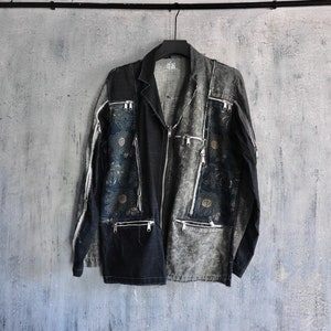 Puede incluir: Una chaqueta vaquera negra y gris con múltiples bolsillos con cremallera y bordados florales. La chaqueta está colgada de una percha negra contra una pared gris.