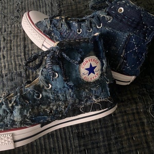 May include: A pair of high-top Converse trainers with a blue denim patchwork design. The trainers have white laces and a white rubber sole. The Converse All Star logo is visible on the side of the trainers.