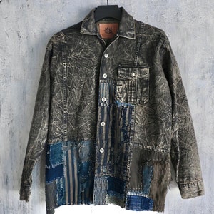 May include: A dark gray denim jacket with a patchwork design. The jacket has a button-up front and long sleeves. The patchwork is made of different shades of blue denim and other fabrics.