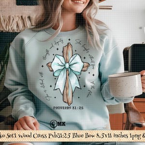 May include: A light blue sweatshirt featuring a wooden cross with a blue bow. The cross has the text "She is clothed with strength and dignity. She laughs without fear of the future." and "Proverbs 31:25".