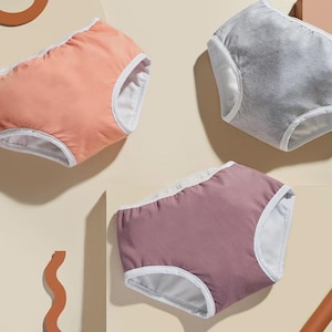 May include: Three pairs of underwear in shades of peach, gray, and mauve, each with white trim. The underwear are arranged on a light beige surface, with a minimalist aesthetic.