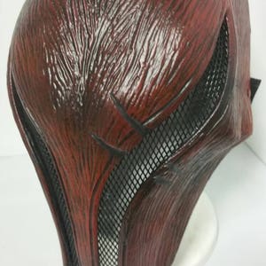 Könnte beinhalten: Eine rote und schwarze Plastikmaske mit einem Netz, das die Augen bedeckt. Die Maske hat eine strukturierte Oberfläche und ein spitzes Kinn.