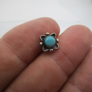 May include: A small silver stud earring featuring a round, bright blue turquoise stone set in the center. The stone is surrounded by a decorative silver frame with four petal-like shapes and small silver spheres at each corner of the setting.