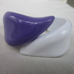 May include: Two plastic bangles, one purple and one white, are shaped like a wave. The bangles are stacked on top of each other.
