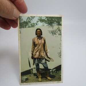 May include: Vintage postcard featuring a Native American man wearing a traditional outfit and holding a hat. The postcard is in color and shows the man standing in front of a white background.
