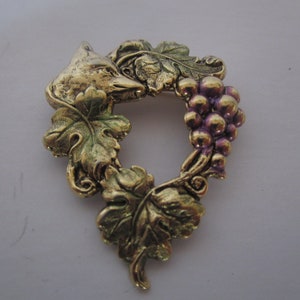 May include: A gold-toned metal brooch featuring a fox's head surrounded by grape leaves and a cluster of purple grapes.