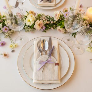 Puede incluir: Elegante mesa con platos blancos, bordes dorados y cubiertos. Un servilletero de punto con una ramita de lavanda y una cinta morada se encuentra en el plato. Copas de vino de cristal y una guirnalda floral completan el conjunto.