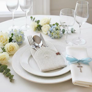 Puede incluir: Elegante mesa con platos blancos, cubiertos y un soporte tejido para utensilios. Una servilleta blanca está atada con una cinta azul y un dije de cruz. La mesa está decorada con rosas blancas y hortensias azules, creando un ambiente sofisticado.