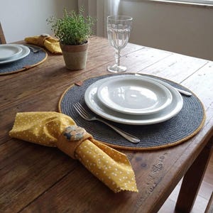 Può includere: Un tavolo di legno rustico apparecchiato per un pasto. Include piatti bianchi, posate, un bicchiere trasparente e un tovagliolo giallo a pois bianchi. Un piccolo vaso di piante e tovagliette grigie con bordi gialli completano l'allestimento.