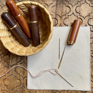 May include: Four wooden needle cases with gold accents are arranged in a woven basket. Three needles are on a beige fabric surface.