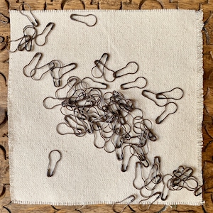 May include: A large pile of antique brass safety pins on a beige fabric surface. The safety pins are all the same size and shape, and they are arranged in a random pattern.
