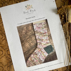May include: A close-up of a colorful sock with a green heel. The sock is on a white sheet of paper with the text "Back Porch Fiber Co. Elevation Socks" and "A simple vanilla sock recipe with a little something extra" printed on it.