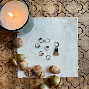 May include: A lit candle in a green glass holder sits next to a collection of small hoop earrings with various gemstone accents. A single earring with a wooden and black resin pendant is also present. Acorns and a beige cloth complete the display.