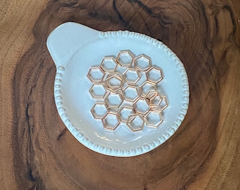 May include: A white ceramic dish with a beaded edge, containing a collection of gold-toned hexagonal metal charms.